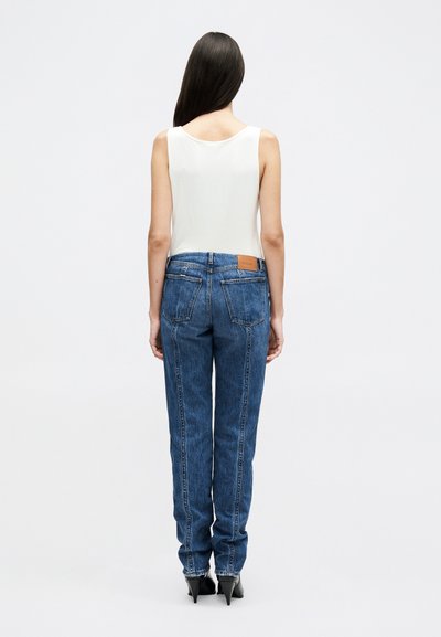 Γυναίκα με μακριά σκούρα μαλλιά που φοράει λευκό αμάνικο τοπ, μπλε τζιν με ψηλή μέση και μαύρα παπούτσια με ψηλό τακούνι, όρθια με την πλάτη προς τα εμπρός.