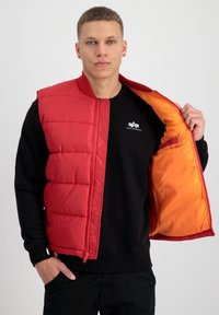 Gilet rembourré rouge avec un design matelassé, fermeture zippée et doublure orange contrastante, porté sur un sweatshirt noir avec logo.