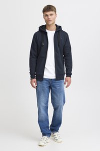 Marineblå lynlås hættejakke med en kængurulomme og snore, parret med en hvid T-shirt, blå jeans og lyse sneakers.