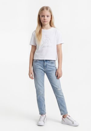 Šviesiaplaukė mergina stovėdama dėvi baltą trumpomis rankovėmis marškinėlį su vienaragio piešiniu, šviesiai mėlynas suplėšytas džinsus ir baltus sportbačius.