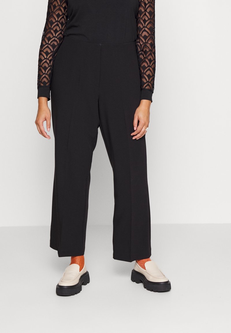 Femme portant un pantalon noir à jambes larges, des manches longues noires transparentes à motifs, des chaussettes orange et des chaussures à plateforme blanches, debout sur fond blanc.