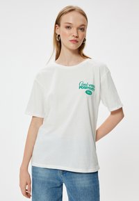 Hvid bomulds-T-shirt med korte ærmer, der har en grøn grafisk tekst "Good Vibes Positivity 90s" på forsiden. Tøjet fremstår afslappet og uformelt.