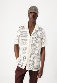 Chemise à manches courtes avec un motif en dentelle de couleur blanc cassé, dotée d'un col, d'une fermeture à boutons sur le devant et de deux poches avant. Complétée par une casquette verte.