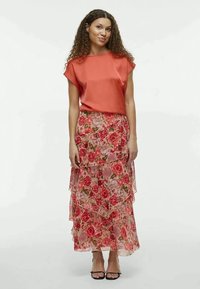 Top in satin corallo abbinato a una maxi gonna a balze con stampa floreale che presenta rose rosa su un tessuto trasparente. La modella indossa sandali neri con cinturini.