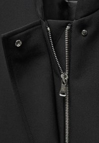 Close-up van een gedeeltelijk open ritssluiting zwarte stoffen jas met zichtbare zilveren rits tanden en drukknopen op de klep.