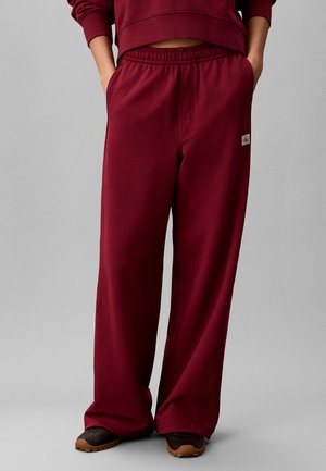 Person iført løse burgundy sweatpants med elastisk talje og sidelommer, kombineret med mørke sko med lysebrune såler, hænderne i lommerne.