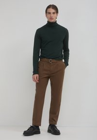 Maglione a collo alto verde scuro e pantaloni marroni con una cintura, abbinati a scarpe nere stringate. Il maglione ha un design aderente.