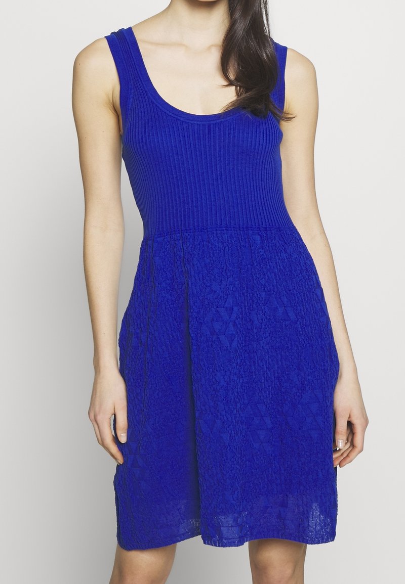 Femme portant une robe bleue sans manches avec un haut côtelé et une jupe texturée sur fond uni.