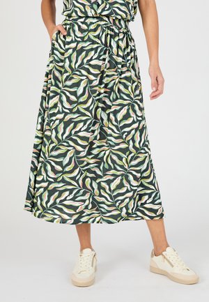 Femme portant une jupe midi verte avec des motifs de feuilles roses et blanches, associée à des baskets beiges à lacets sur un fond uni.