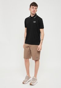 Polo shirt nera con dettagli bianchi sul collo, pantaloni corti beige e sneakers chiare con logo. Design semplice e casual, maniche corte e vestibilità aderente.