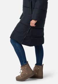 Lang, mørk pufferjakke med horisontale sømme, blå jeans og brune støvler til midt på læggen med tekstureret sål og snørebånd.
