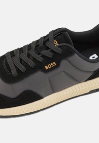 Sneaker nera e grigia con un mix di suede e materiali lisci, suola in gomma crema a contrasto e logo prominente sul lato.