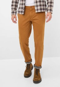 Braune Baumwoll-Chinos mit schmaler Passform, ausgestattet mit einem Knopfverschluss und aufgerollten Bündchen über braunen Wildlederstiefeln mit schwarzen Sohlen.