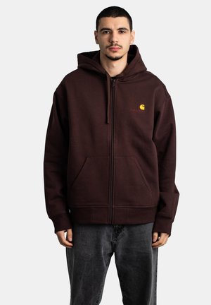 Joven con cabello corto que lleva una sudadera con capucha marrón oscuro con cremallera y un logo amarillo, y vaqueros gris oscuro, de pie frente a un fondo liso.