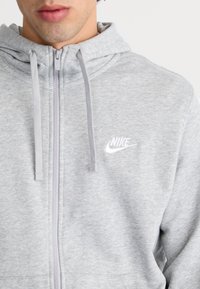 Felpa grigia con zip realizzata in un tessuto morbido, dotata di tasca a marsupio, con un piccolo logo Nike bianco sul lato sinistro del petto e cappuccio con cordino.