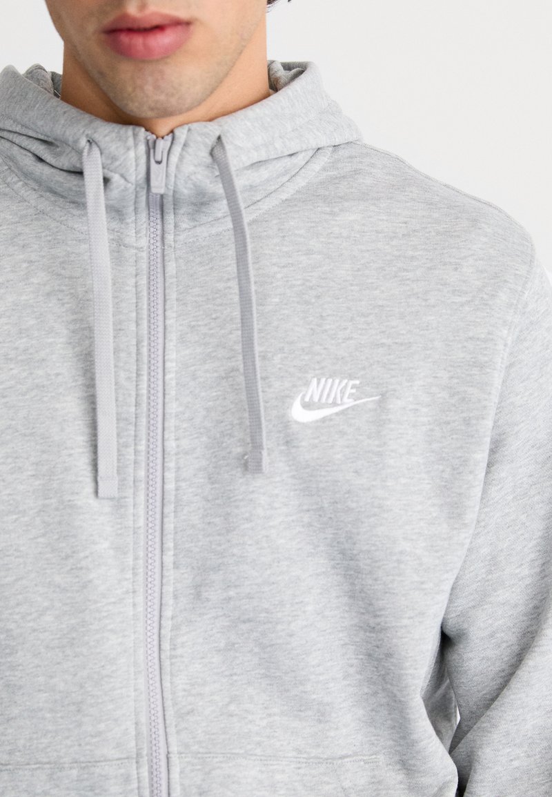 Sudadera con cremallera gris hecha de una tela suave, con un bolsillo canguro, que presenta un pequeño logo blanco de Nike en el lado izquierdo del pecho y una capucha con cordón.