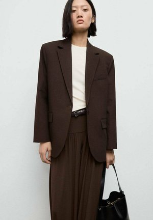 Blazer marrone su misura con una tasca, indossato sopra una maglietta chiara e pantaloni marroni abbinati. Tiene una borsa nera. Tessuto liscio, design strutturato.