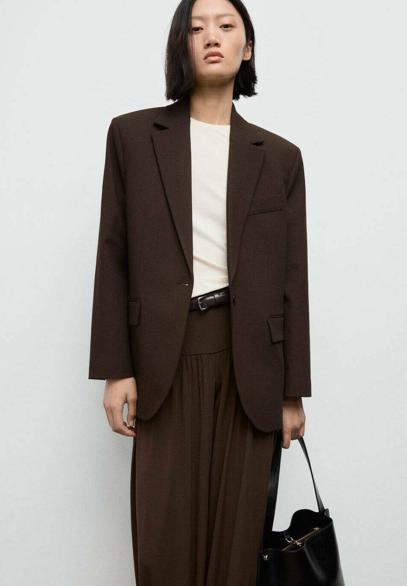 Blazer marrone su misura con una tasca, indossato sopra una maglietta chiara e pantaloni marroni abbinati. Tiene una borsa nera. Tessuto liscio, design strutturato.