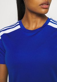 Camiseta atlética azul de manga corta, con cuello redondo, que presenta rayas blancas a lo largo de los hombros. Textura suave, diseño ajustado.