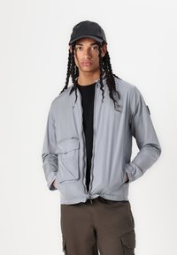Šviesiai pilka užtraukiama striukė su priekinėmis kišenėmis ir elastingais rankogaliais, derinama su juoda kepure ir juodu marškinėliais po ja. Modelis vilki khaki spalvos kelnes.