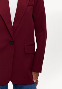 Bordeaux blazer van gladde stof, met een enkele zwarte knop, een borstzak en slanke mouwen, gecombineerd met blauwe denim.