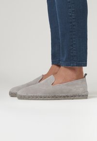 Graue Slipper aus Wildleder mit einer gewebten Jutesohle, runder Zehenpartie und ohne sichtbare Beschläge. Strukturierte Oberfläche mit weichem Finish.