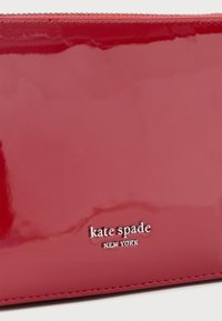 Fényes piros bőr pénztárca ezüst "kate spade NEW YORK" logóval, amely a alsó szél közelében középen helyezkedik el, fehér háttéren.