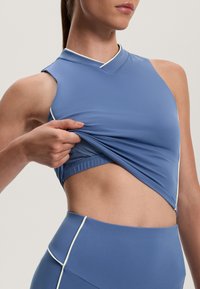 Modra športna majica brez rokavov, z rahlim V-izrezom in belimi robovi; material se zdi elastičen in prilega.