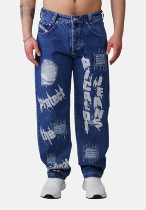 Blue denim jeans with white graphic text reading "Protect the original" and "DLTAP JEANS," worn with white shoes and a tattooed arm visible.