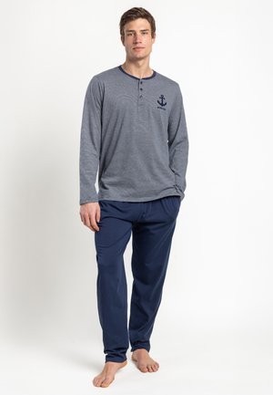 Joven de pie descalzo vistiendo una camisa henley de manga larga gris con logo de ancla y pantalones azul marino sobre fondo blanco.
