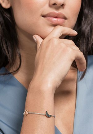 Woman’s wrist with silver chain bracelet featuring a crescent moon charm with dark stones and a circular gold charm, hand resting near face.