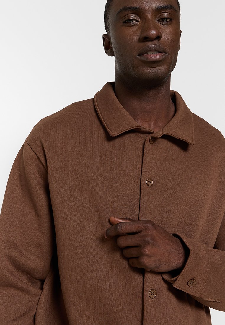 Chemise marron à boutons avec un col évasé, en tissu texturé, à manches longues, comportant deux boutons aux poignets et une coupe décontractée.