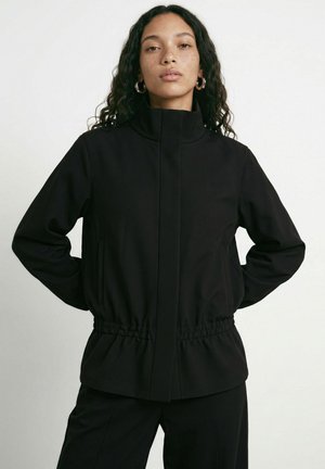 Jeune femme aux longs cheveux bouclés portant une veste noire à col montant et taille élastique, debout, les mains derrière le dos.