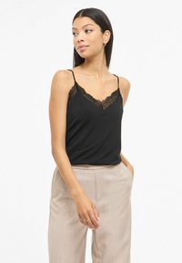 Czarny top na cienkich ramiączkach z koronkową lamówką, z regulowanymi paskami i gładką, miękką fakturą. W połączeniu z jasnymi spodniami.