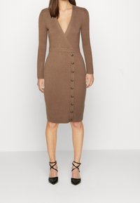 Donna che indossa un abito marrone a maglia a coste, aderente, con maniche lunghe, scollatura a V profonda, dettagli con bottoni sul davanti e tacchi neri a punta con cinturini.
