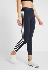 Navy leggings with white three-stripe design on sides, high waistband, made of stretchy material, paired with light gray athletic shoes.