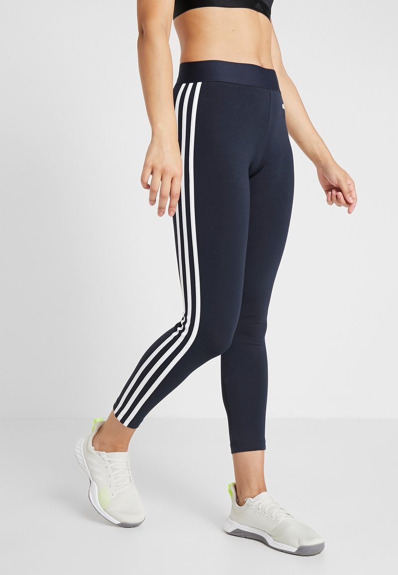 Navy leggings with white three-stripe design on sides, high waistband, made of stretchy material, paired with light gray athletic shoes.