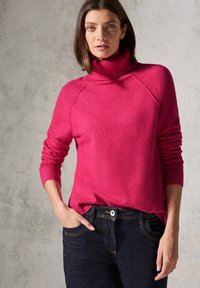 Fuchsia Rollkragenpullover mit Rippdetails, weicher Textur und Raglanärmel, kombiniert mit dunkelblauen Jeans aus Denim mit kontrastierenden Nähten.