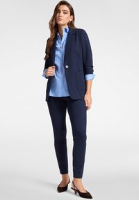Blazer navy con un solo botón, combinado con una camisa a rayas azul claro y pantalones navy a juego. Corte ajustado y textura de tela suave.