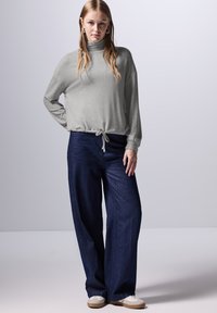 Lichtgrijze, lange mouwen coltrui met een trekkoord in de taille, gecombineerd met wijde donkerblauwe spijkerbroeken en witte sneakers.