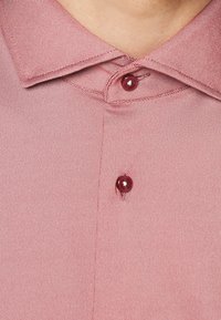 Primo piano di un colletto di camicia rosa strutturato con due bottoni color bordeaux abbottonati attorno al collo e alla parte superiore del torace di una persona.