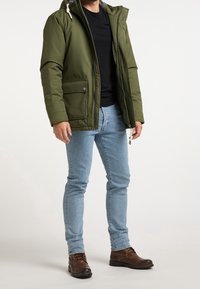 Grüne isolierte Jacke mit Kapuze, elastischen Bündchen und Reißverschluss-/Knopfverschluss. Getragen mit einem schwarzen Hemd, blauen Jeans und braunen Stiefeln.