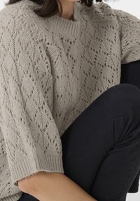 Personne portant un pull en tricot gris clair avec des motifs en losange et un pantalon sombre, assise avec un genou plié près du corps.