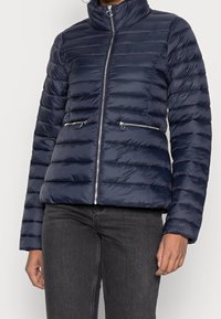 Giacca puffer blu navy con design trapuntato orizzontale, collo alto, zip frontale e due tasche laterali con zip. Texture liscia e lucida.