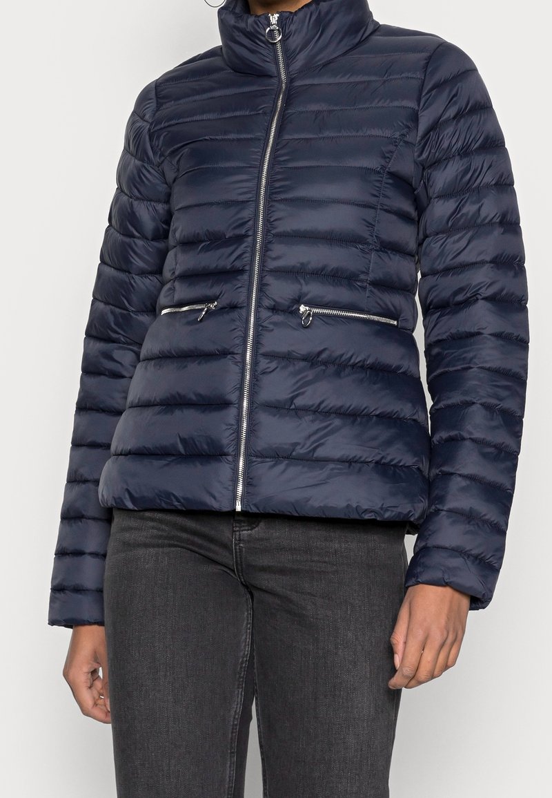Giacca puffer blu navy con design trapuntato orizzontale, collo alto, zip frontale e due tasche laterali con zip. Texture liscia e lucida.