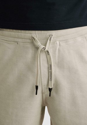 Béžové rebrované nohavice s šnúrkou zviazanou do mašle, čierne konce a nápis „PRIME LEGEND“ na šnúrkach, nosené s čiernou košeľou.