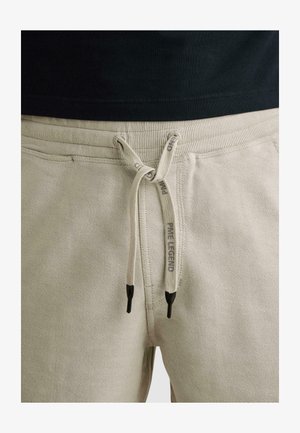 Béžové rebrované nohavice s šnúrkou zviazanou do mašle, čierne konce a nápis „PRIME LEGEND“ na šnúrkach, nosené s čiernou košeľou.