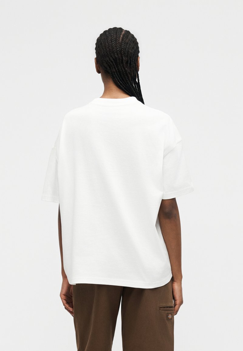 Persona con capelli intrecciati, indossa una camicia bianca a maniche corte e pantaloni marroni, vista da dietro su uno sfondo semplice.