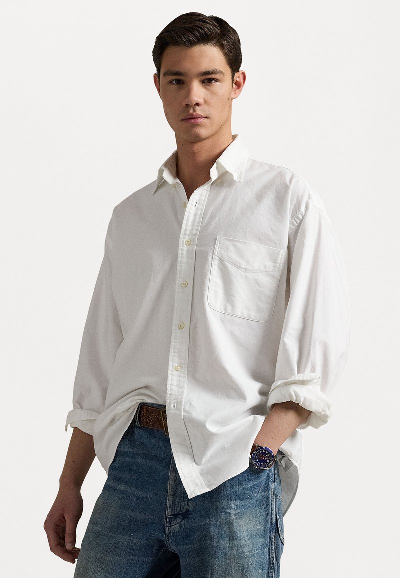 Camisa blanca de botones con mangas largas y un único bolsillo en el pecho, combinada con jeans de mezclilla azules. Reloj circular en la muñeca.