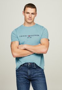 Camiseta de algodón de color azul claro con mangas cortas y el logo "TOMMY HILFIGER EST. 1985" en azul oscuro, combinada con jeans de color azul oscuro.